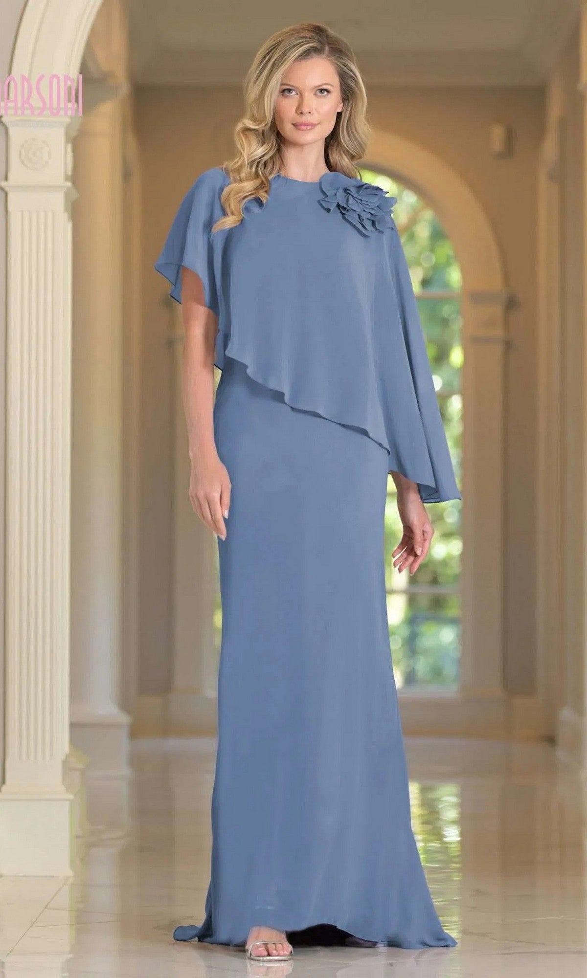 A woman stands indoors between white columns, gracefully posing in the Marsoni MV1347 long formal dress in slate blue with floral applique, chiffon fabric, and an asymmetrical capelet sleeve.